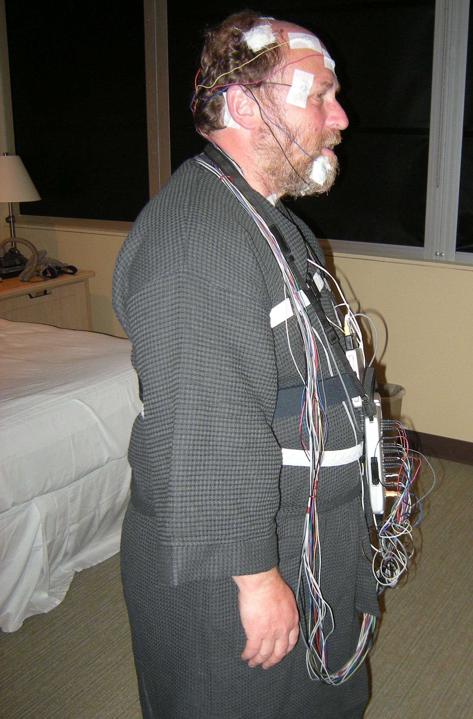 A man standing in a bedroom with electrodes taped to his head and face and a bundle of coloured wires running to a recorder at his waist.