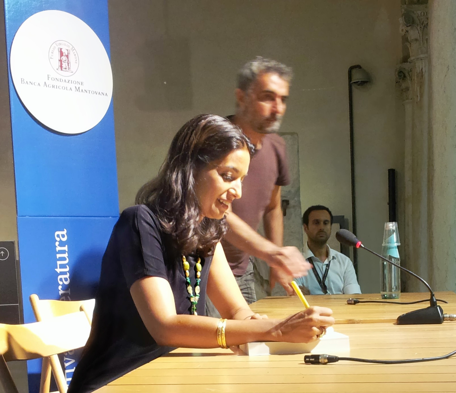 A woman signing a book at a festival table, a microphone and a festival banner beside her.