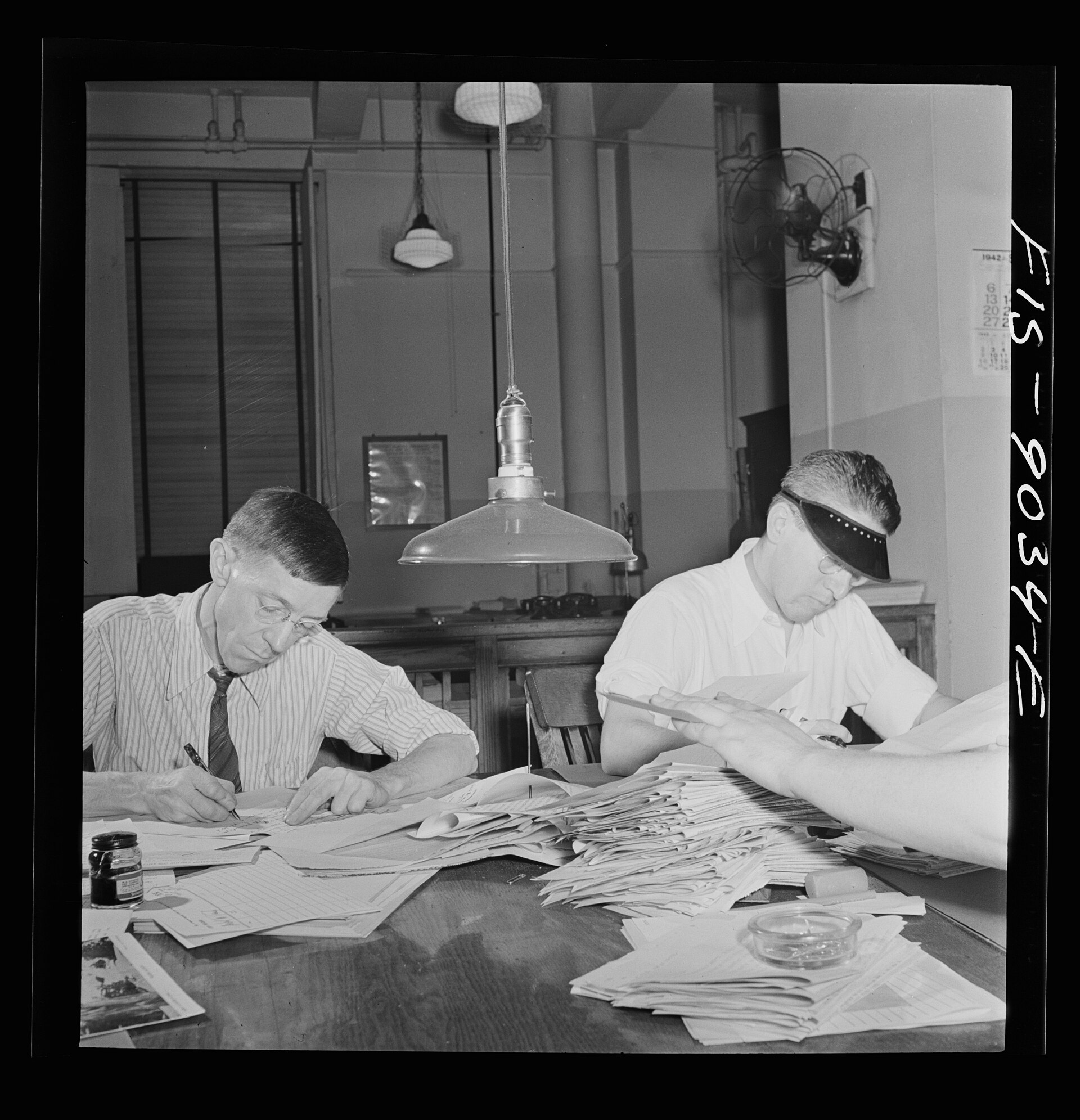 Two men in shirtsleeves bent over paper at a newspaper copy desk under a hanging lamp, with a hand reaching in from the right holding more pages.