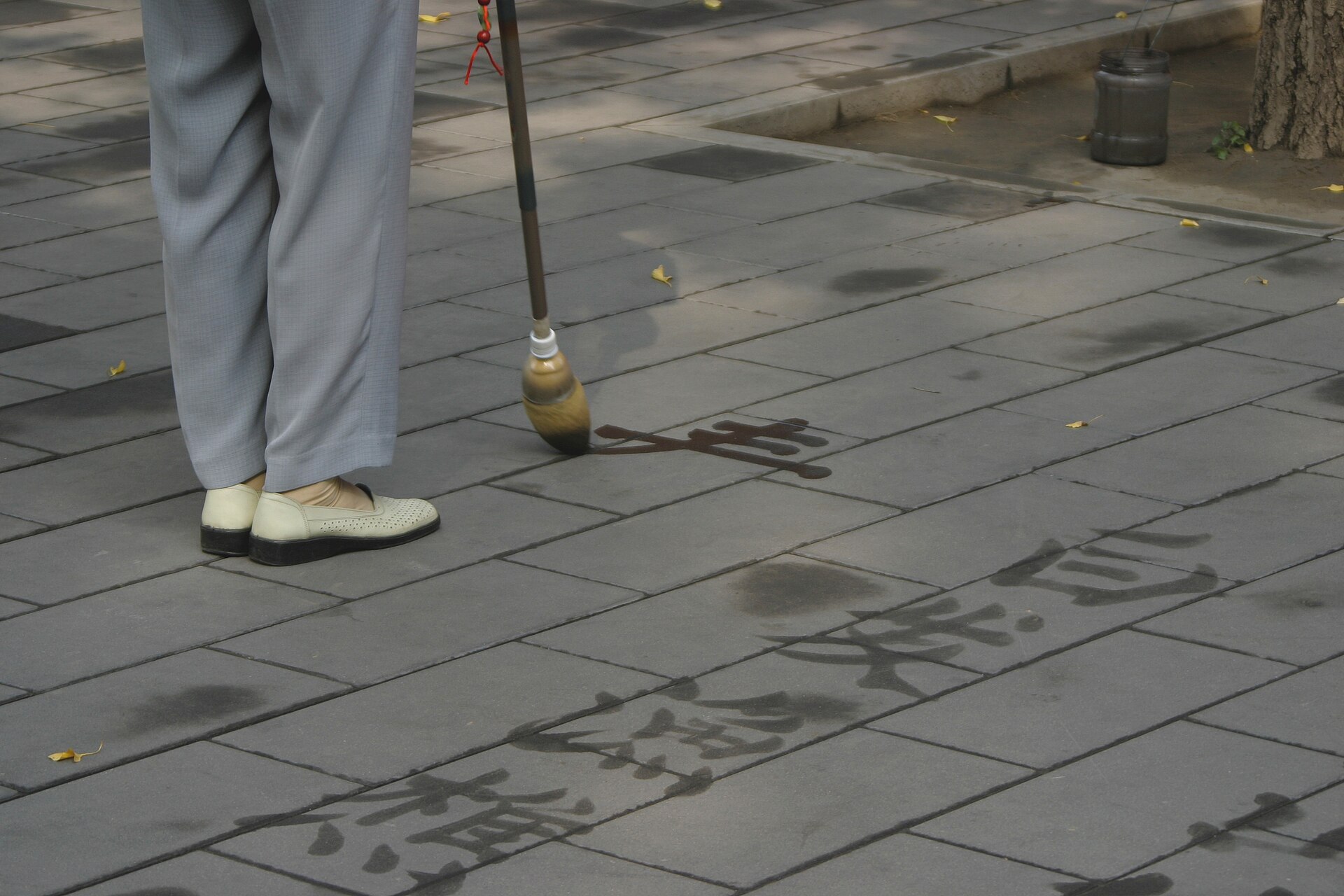 A person writing large characters on paving stones with a long brush dipped in water.