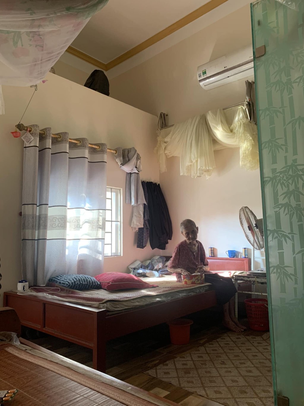 Grandma sitting on the bed in a sunlit room, a bowl in her hands; beside her a hanging mosquito net, the curtains and an electric fan