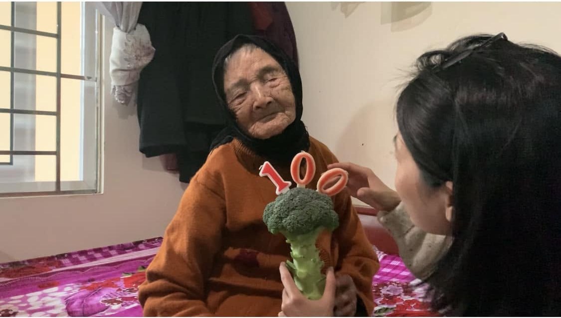 Thom’s grandmother in a black headscarf and an orange jumper, sitting on a bed with a floral quilt, holding a head of broccoli with candles reading 100 in place of a birthday cake; a grandchild leans in from the right