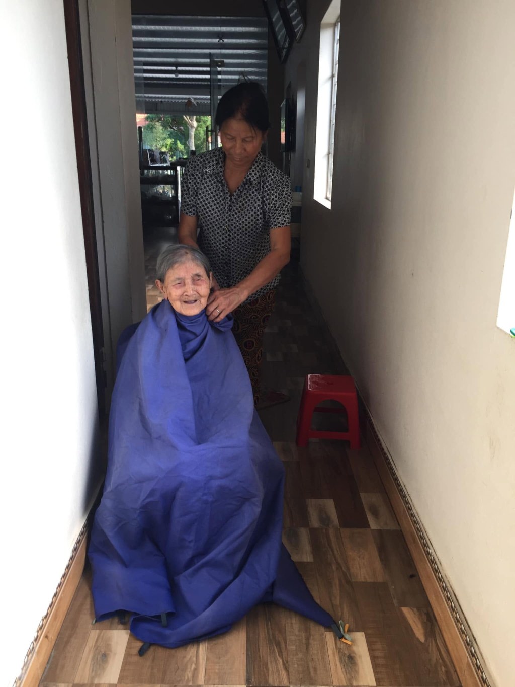 Grandma sitting in a doorway, wrapped from the shoulders down in a blue hairdressing cape, smiling; behind her, bác Xoan stands cutting her hair. A corridor of wood-effect tiles, a red plastic stool beside them