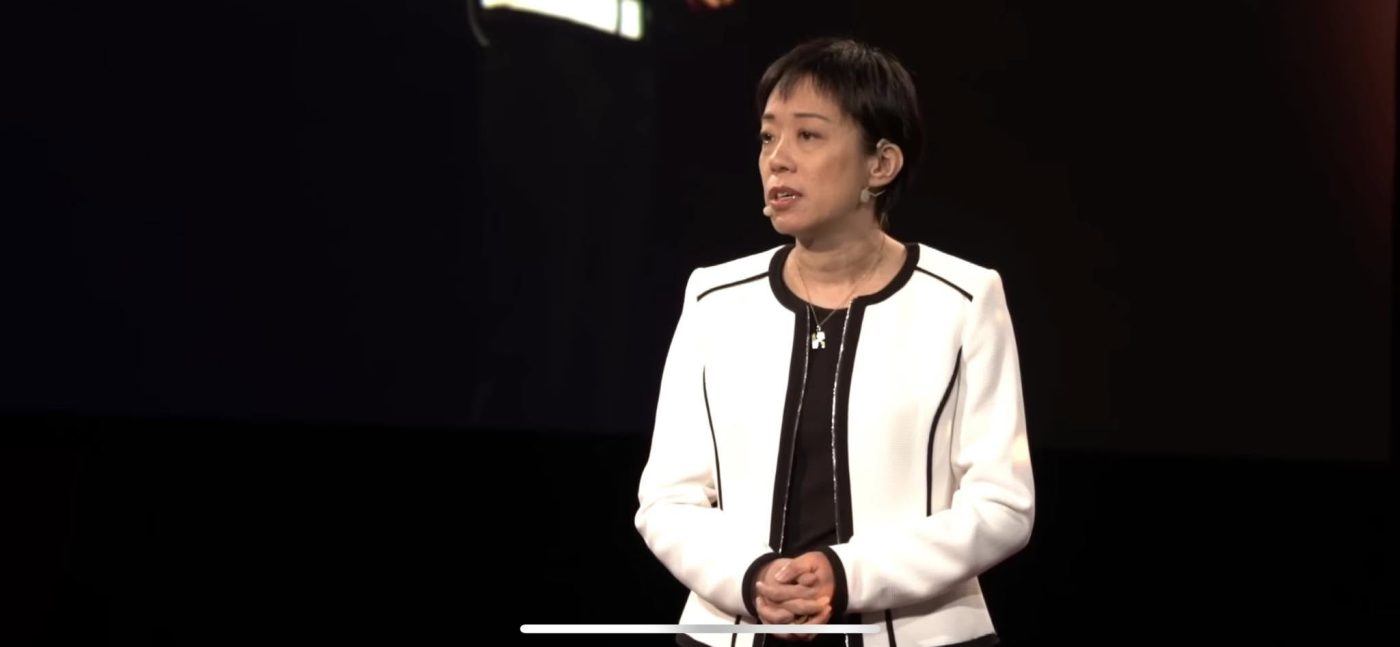 May Chen in a white jacket with black trim, wearing a headset microphone, standing with her hands clasped as she speaks on a TEDx stage against a dark backdrop