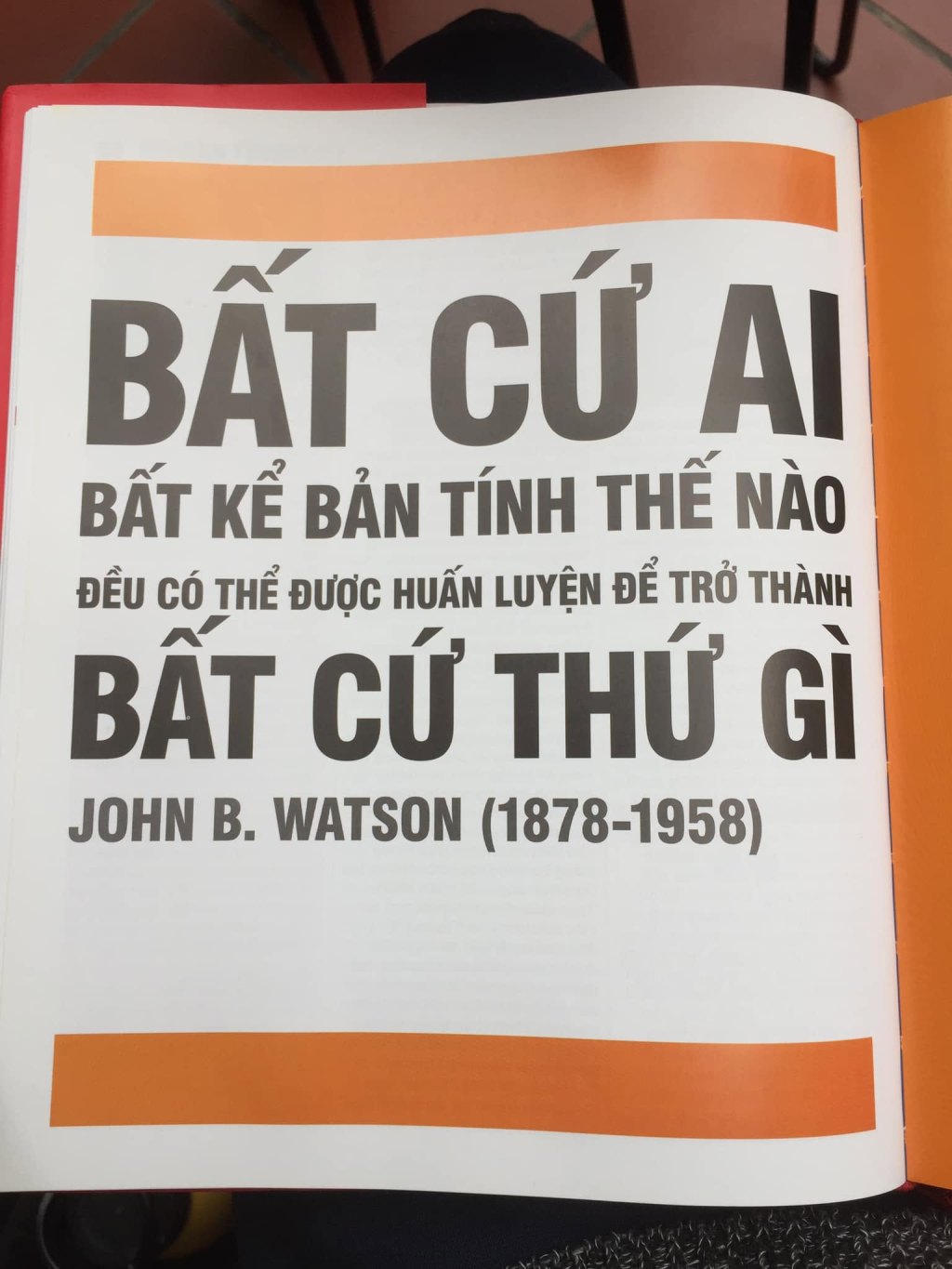A book page set in large grey capitals on white, with orange bands top and bottom, reading in Vietnamese “ANYONE, WHATEVER THEIR NATURE, CAN BE TRAINED TO BECOME ANYTHING”, and beneath it “JOHN B. WATSON (1878-1958)”