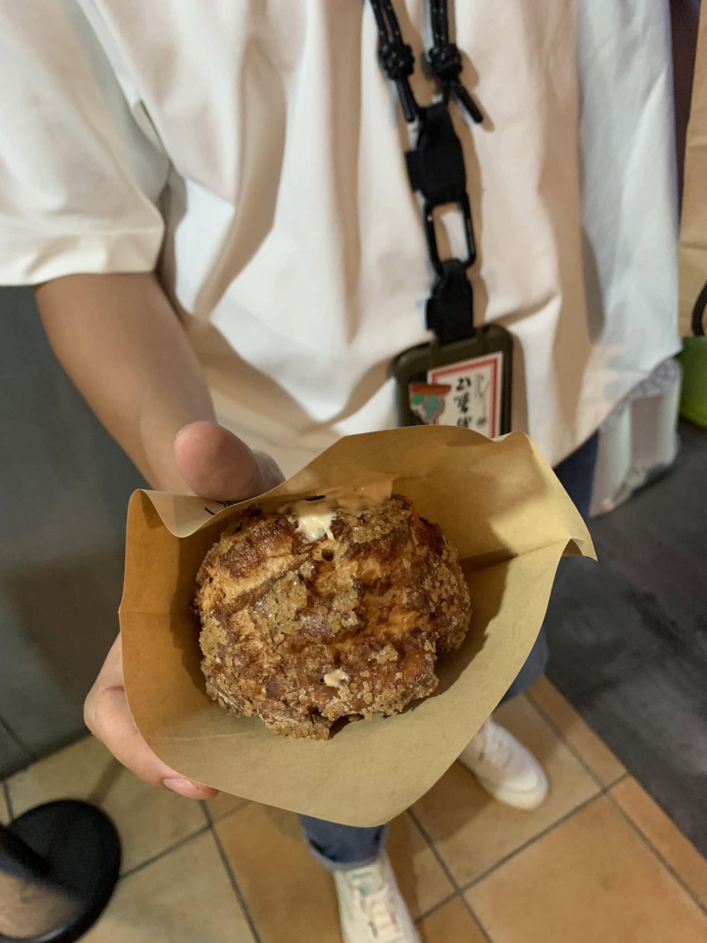 A hand holding out a round bun with a cracked, crisp sugar crust, wrapped in brown paper, tea cream spilling from the top; behind, a person in a white shirt with a lanyard, standing on a tiled floor