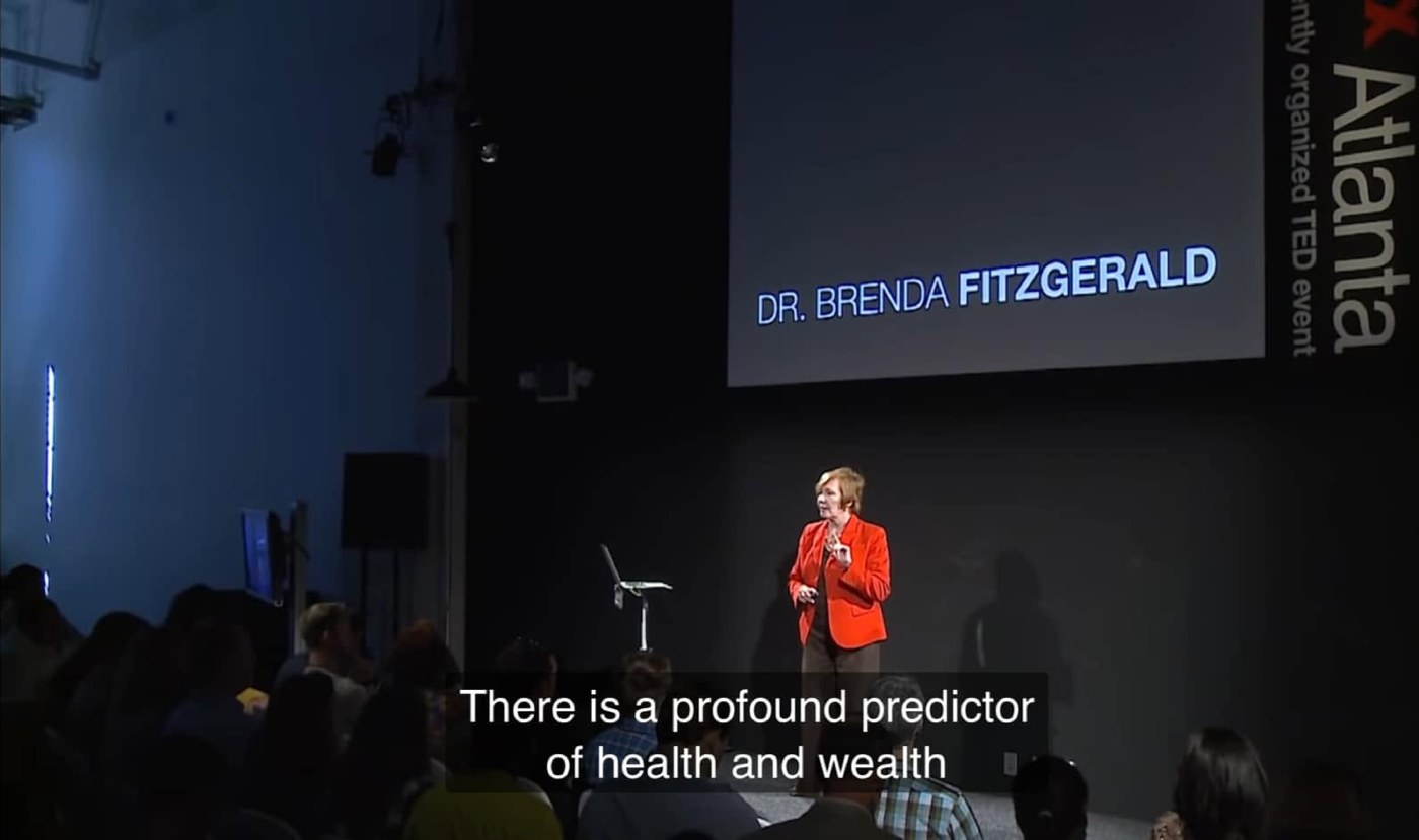 Dr Brenda Fitzgerald in a red jacket speaking on the TEDxAtlanta stage, the screen behind her showing her name; the subtitle at the bottom reads “There is a profound predictor of health and wealth”