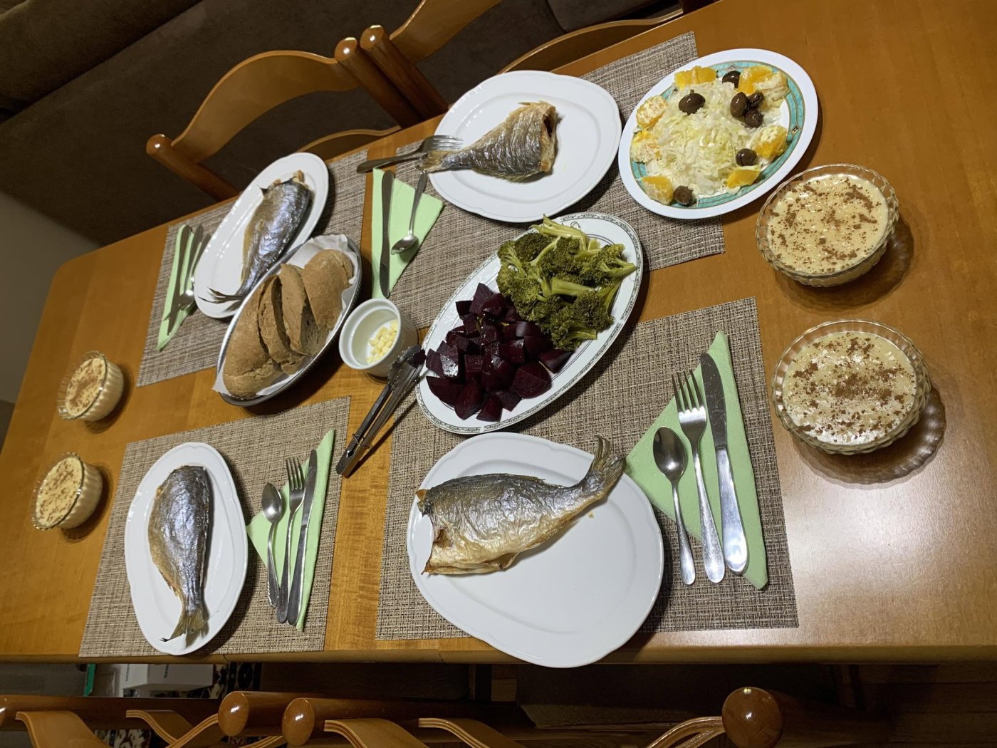 Looking down at a laid wooden dining table: three plates of whole grilled fish, a dish of broccoli and diced beetroot, a cabbage salad with orange and olives, a basket of brown bread, and several bowls of milk pudding dusted with cinnamon