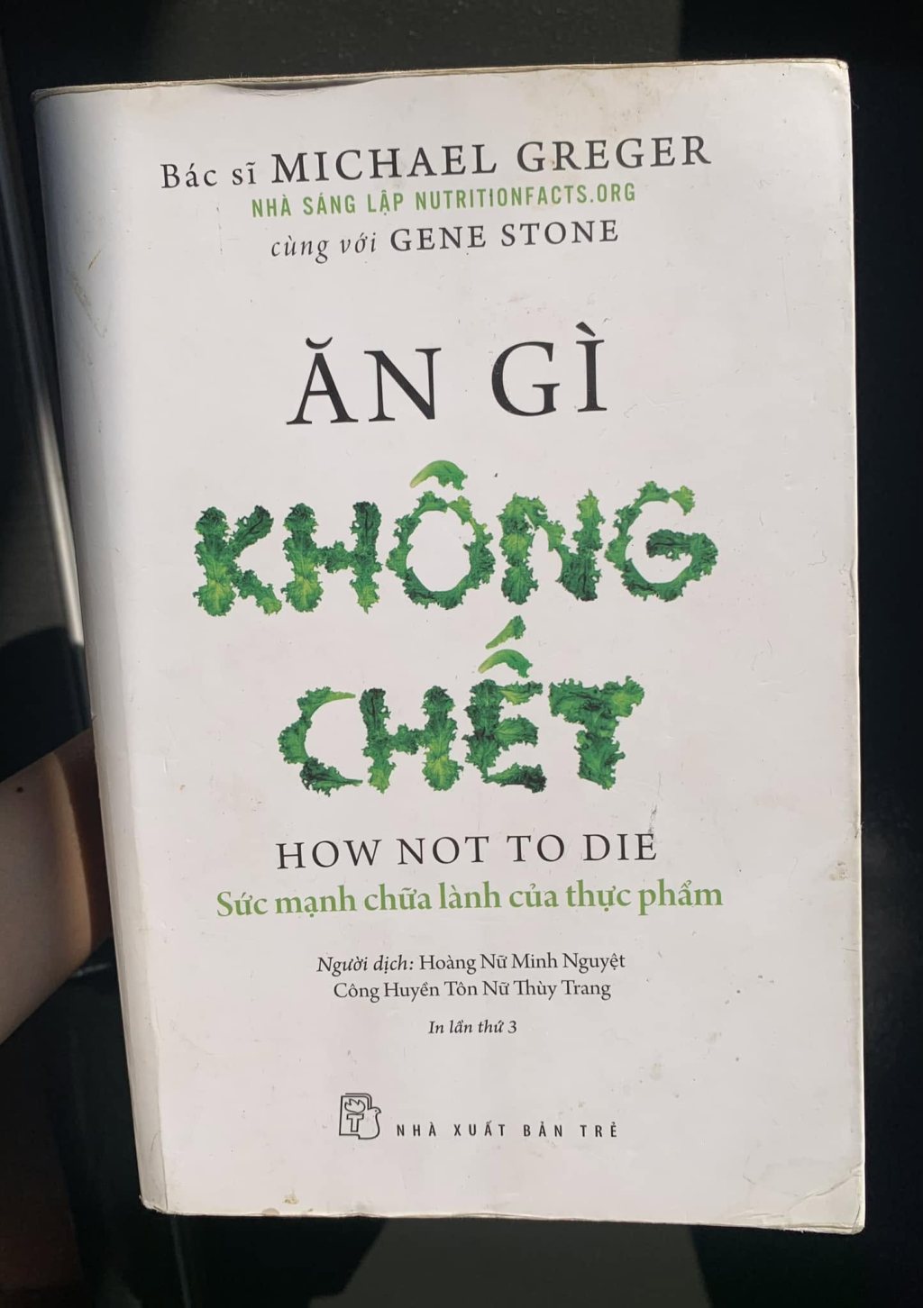 The title page of a book on white: the authors “Bác sĩ MICHAEL GREGER, founder of NUTRITIONFACTS.ORG, with GENE STONE”, the Vietnamese title “ĂN GÌ KHÔNG CHẾT” with the words “KHÔNG CHẾT” made out of green kale leaves, and below it “HOW NOT TO DIE” and the publisher Nhà xuất bản Trẻ
