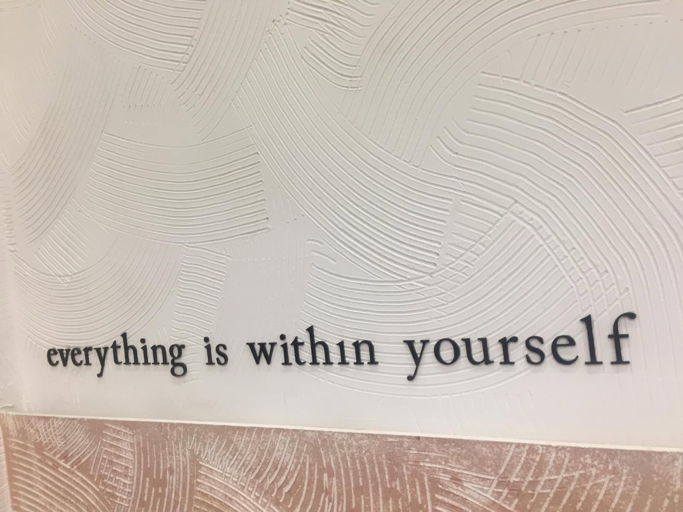 A white plaster wall in a café, its surface combed into sweeping curves, with the words “everything is within yourself” mounted on it in dark letters; below runs a terracotta band combed in the same way