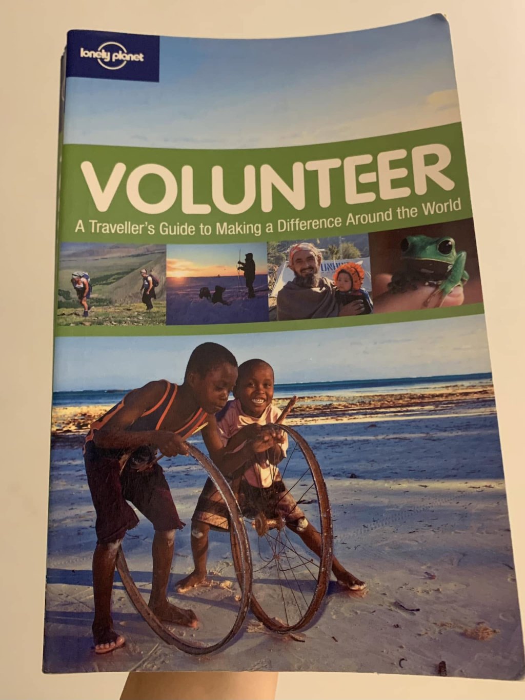 The cover of a worn Lonely Planet book, the word “VOLUNTEER” in large white letters on a green band; below it a photograph of two boys rolling a bicycle rim along a white sand beach