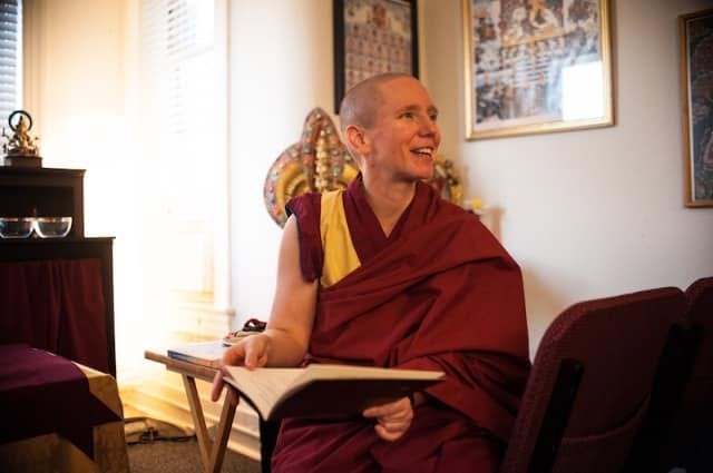 Gen Kelsang Nyema in maroon and yellow robes, smiling brightly as she sits by a window with an open book on her lap, Buddhist paintings hanging on the wall behind her
