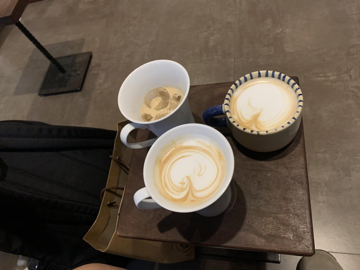 Looking down at three cups of coffee on a small wooden table: two still carry latte art on the milk foam, one of them a white cup with blue striped trim; the third is nearly finished, with only ice and a little milk left