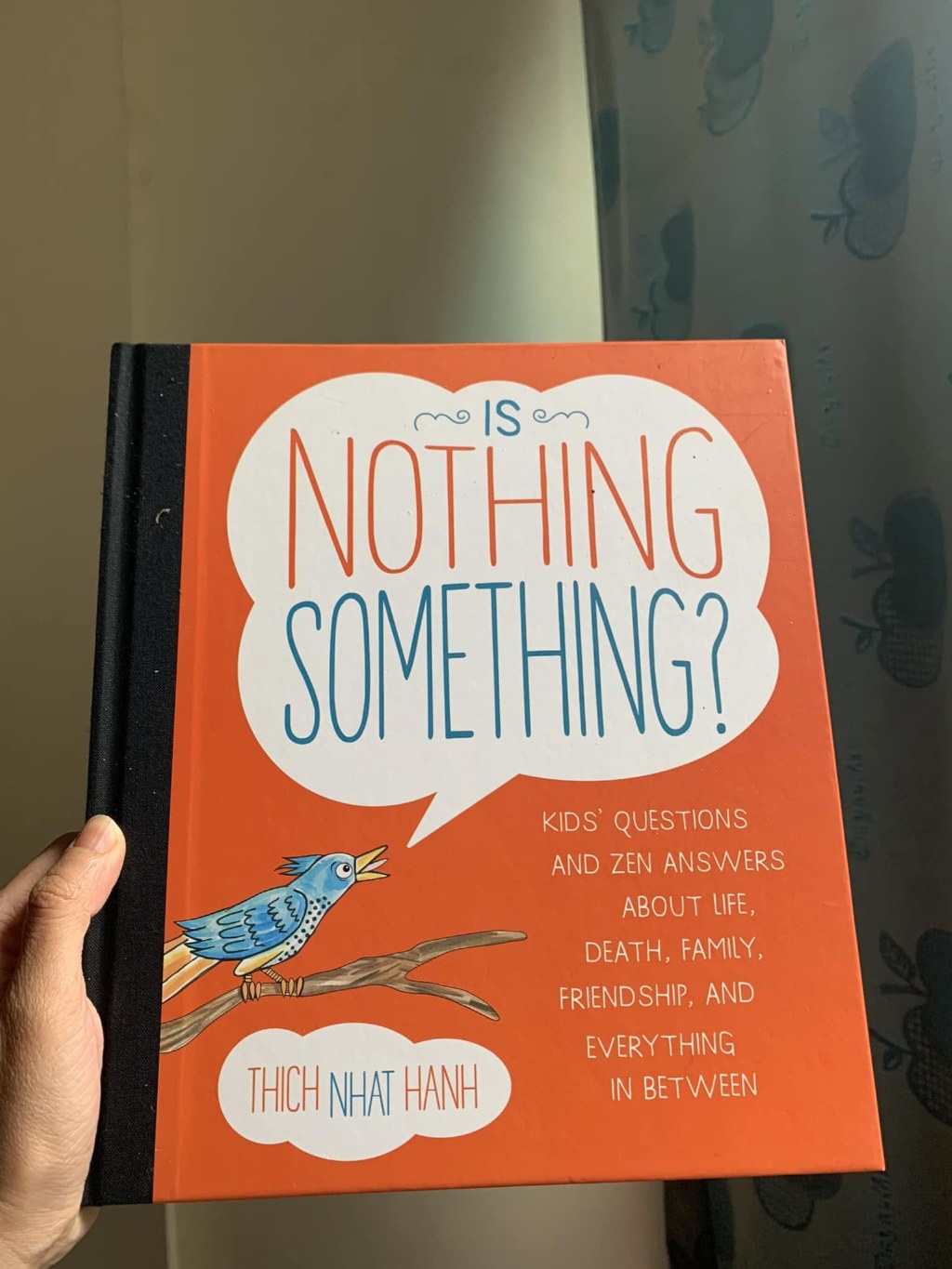 A hand holding the orange-covered book “Is Nothing Something?” by Thích Nhất Hạnh: the title sits in a white speech bubble, and below it a blue bird sings on a branch