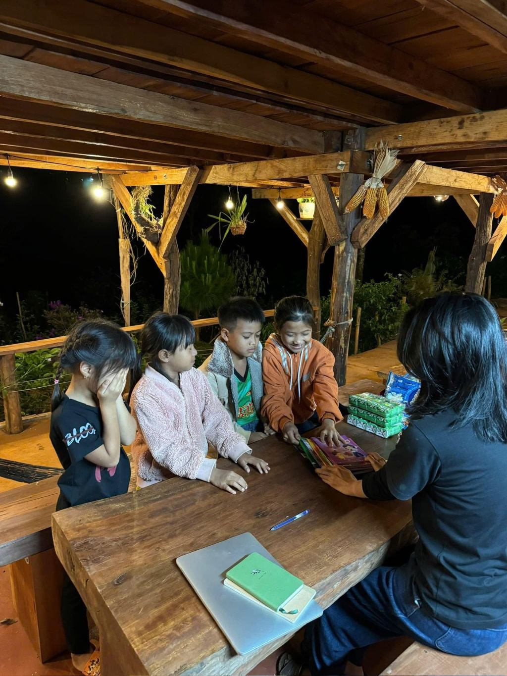 Four children sitting round a wooden table under a veranda at night, a stack of books and gift boxes in front of them; Thom sits opposite with his back to the camera
