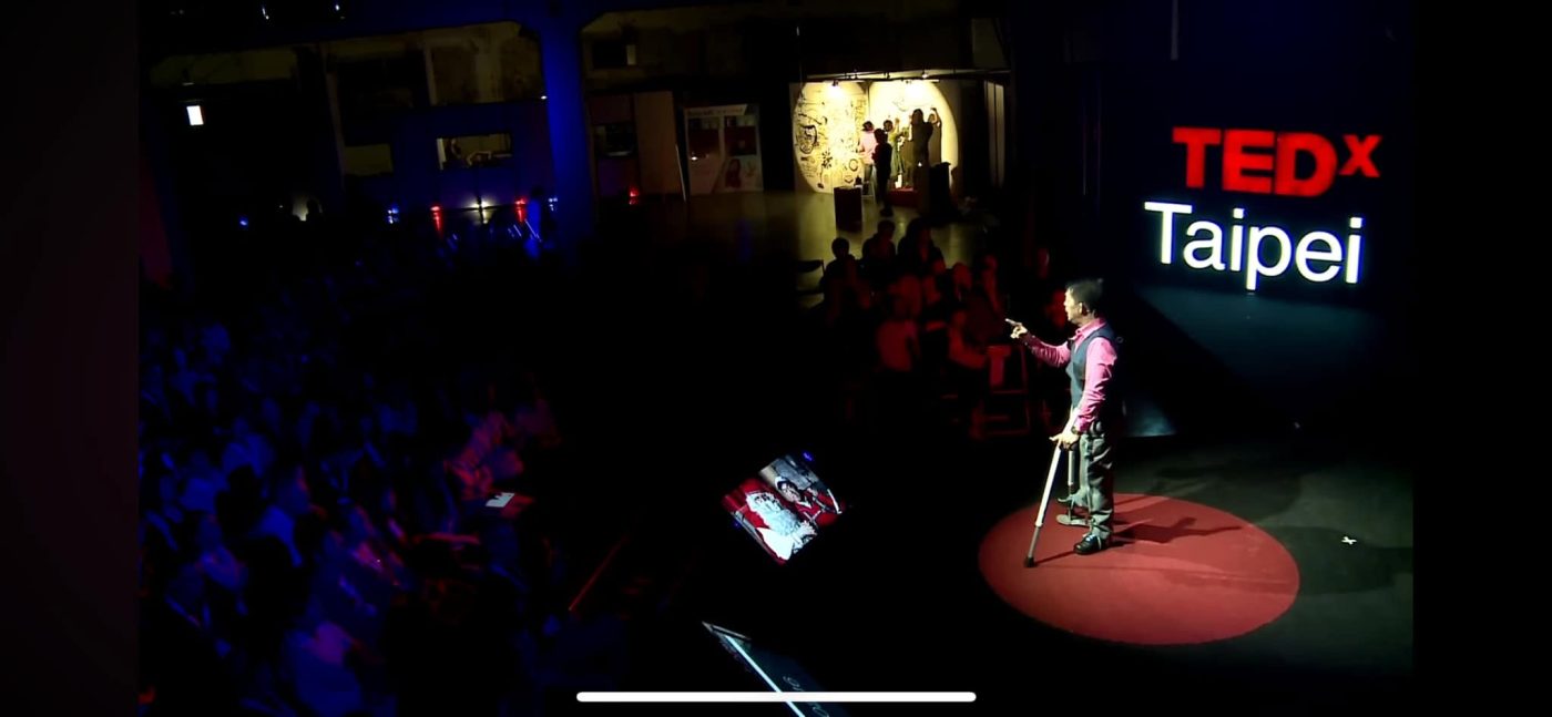 Logan Hsu standing with a crutch on the red circle at the centre of the TEDxTaipei stage, pointing towards the audience, the lit TEDx Taipei sign behind him