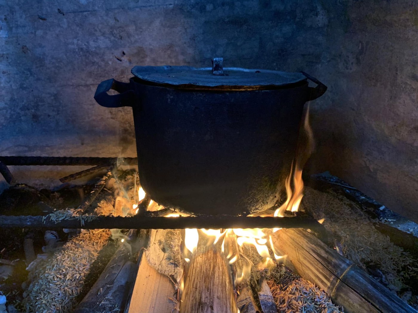 A soot-blackened aluminium pot with its lid on, standing over a wood fire in a whitewashed kitchen; flames licking around split logs, rice husks scattered on the floor