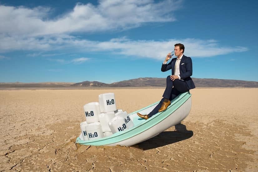 A staged photograph: a man in a navy suit sits on the prow of a small boat stranded on a cracked, dried-up lake bed, drinking from a glass; the boat is loaded with white jerrycans marked H2O