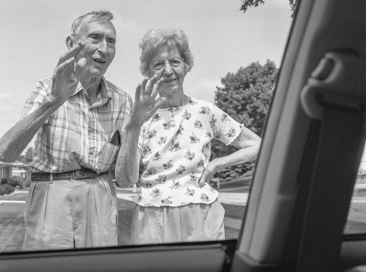 A black-and-white photograph taken through a rolled-down car window: an elderly man in a checked shirt and an elderly woman in a floral blouse standing side by side on the lawn in front of their house, both raising a hand to wave