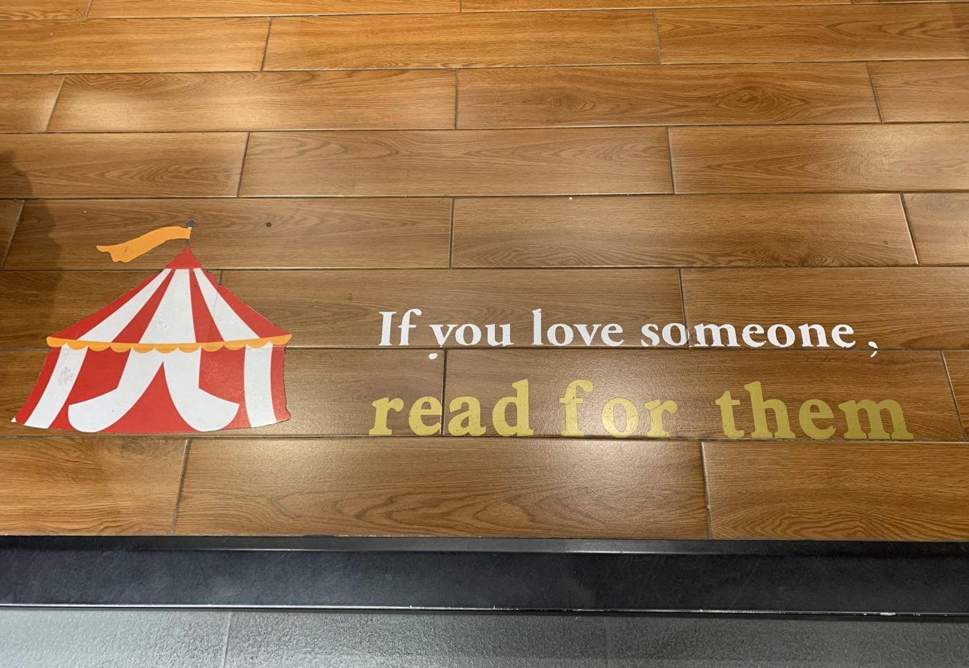 The wooden floor of a bookshop, with a red and white circus tent painted beside the words If you love someone, read for them