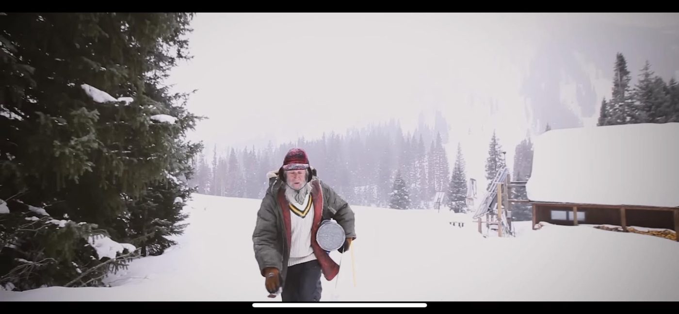 An older man in a red striped woollen hat and a thick coat, carrying a metal pan, walking across white snow among pine trees; to the right is a wooden cabin with a snow-covered roof