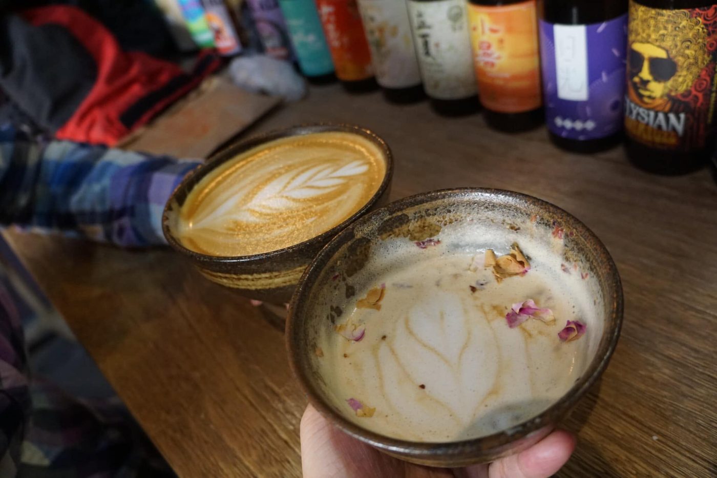 Two brown-glazed ceramic bowls of milk coffee with leaf patterns drawn in the foam; the nearer one is held in a hand and scattered with dried rose petals, on a wooden counter with a row of bottles behind