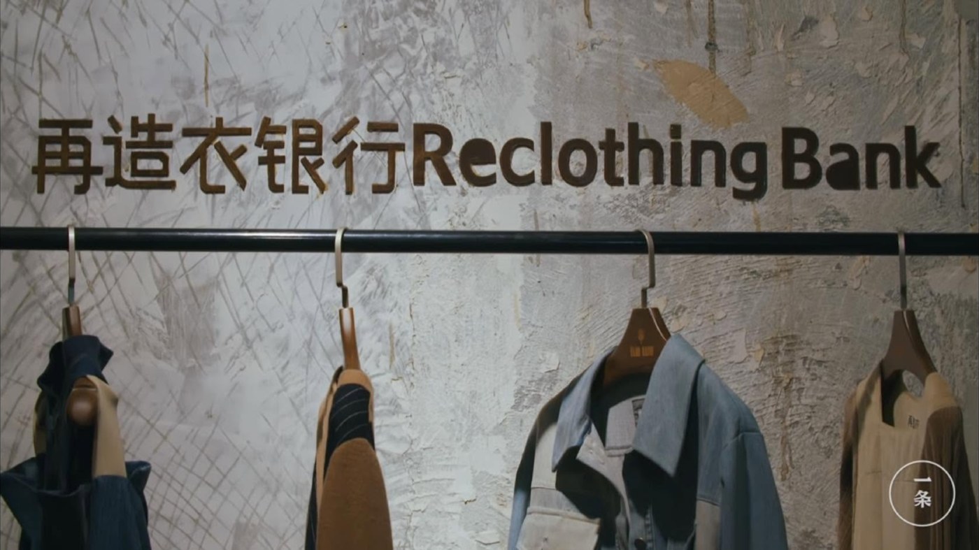 The 再造衣银行 Reclothing Bank sign on a weathered plaster wall in Shanghai, with a rail of garments remade from old cloth hanging beneath it