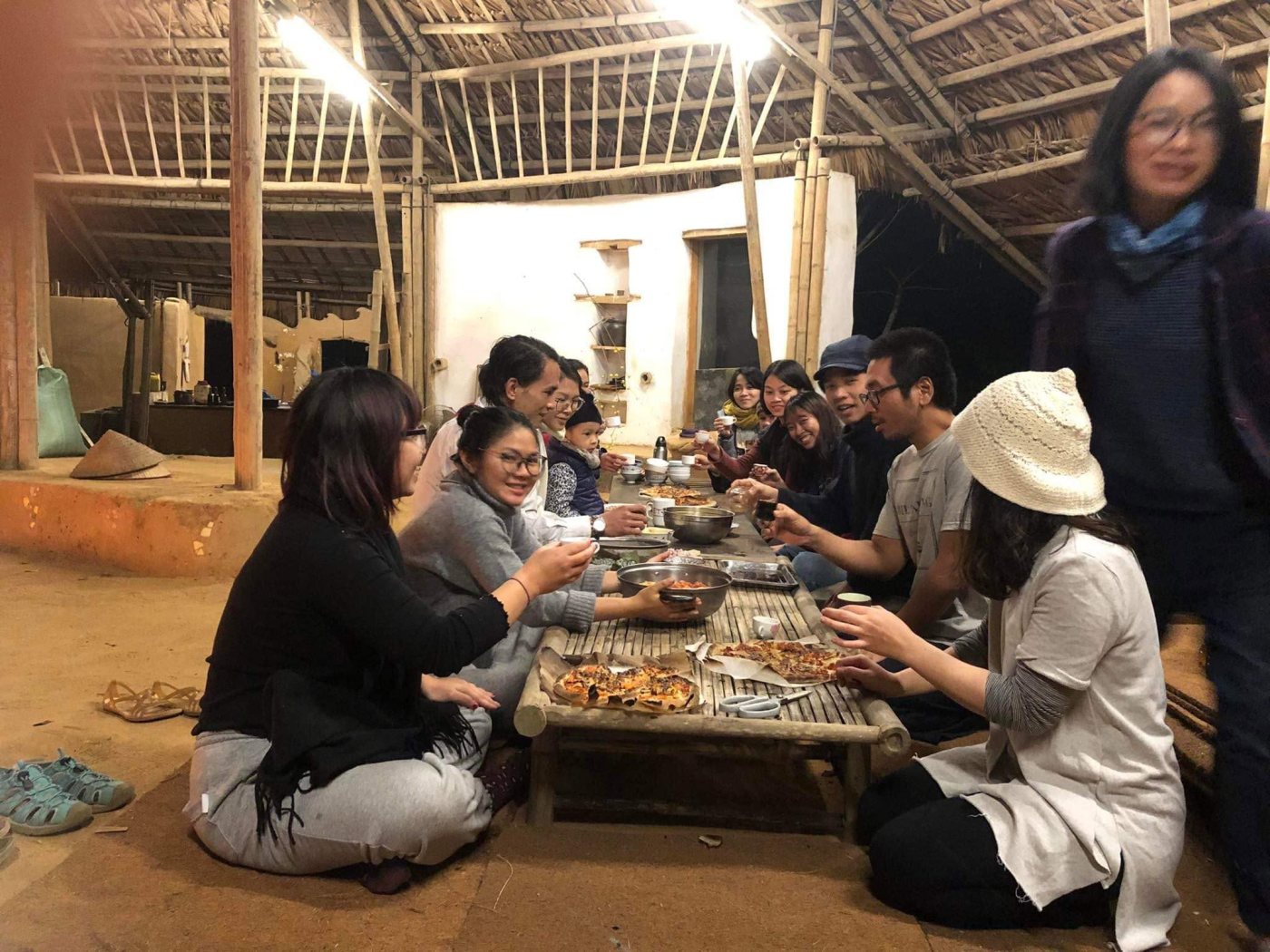 About ten people sitting on the earth floor around a low bamboo table at night, inside an earth-walled building with a thatched roof; home-made pizzas lie on the table and several people are raising their cups