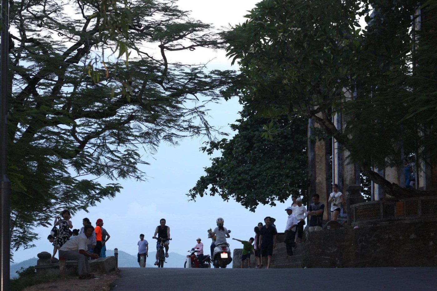 The slope up to Thiên Mụ pagoda in the late afternoon: people walking, a cyclist and a few motorbikes on the road, broad trees closing overhead on both sides, and the river and hazy hills beyond