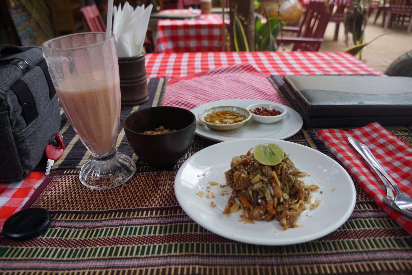 A meal on a table laid with a red-and-white checked cloth and a woven mat: a tall glass of pink smoothie, a plate of salad with a wedge of lime, a wooden bowl of roasted peanuts, two small dishes of dressing and chilli flakes; a camera bag at the left