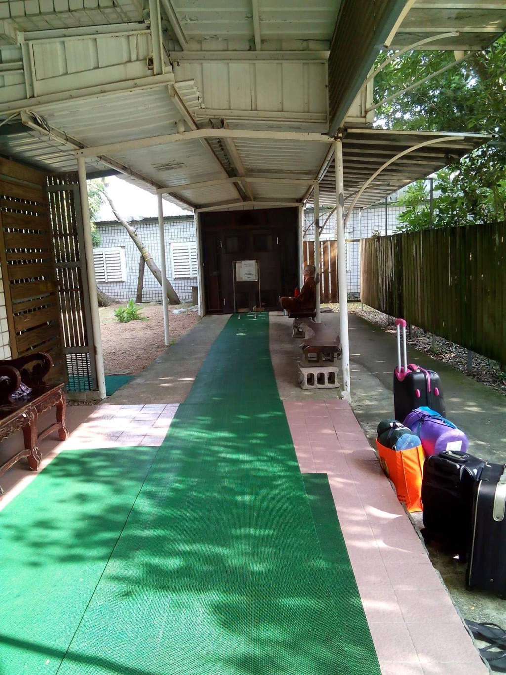 A covered walkway leading to the Vipassana meditation hall: a green mat runs the length of it to a dark wooden door, a monk in maroon robes sits on a bench beside the door, and suitcases and bags belonging to the students are lined up on the right