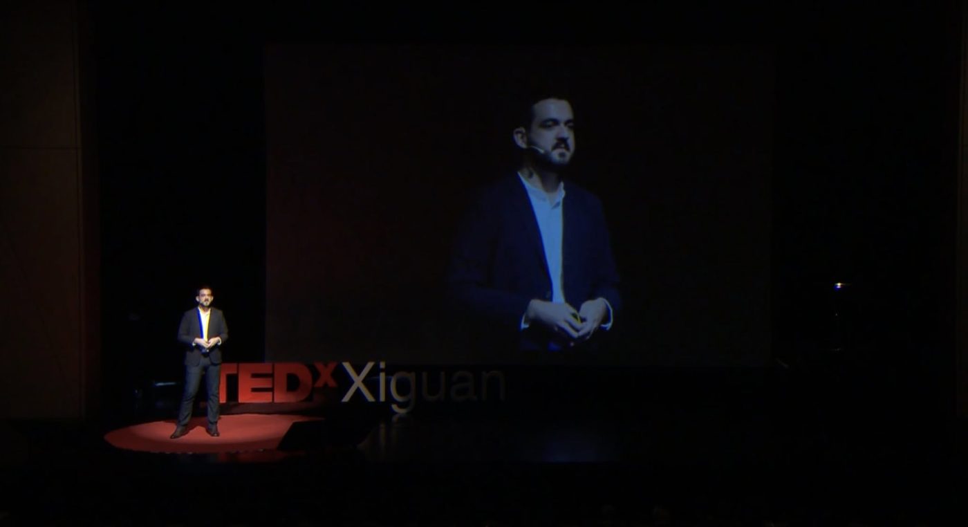 Shane Leaning alone on the TEDxXiguan stage, a large screen behind him showing a close-up of his face