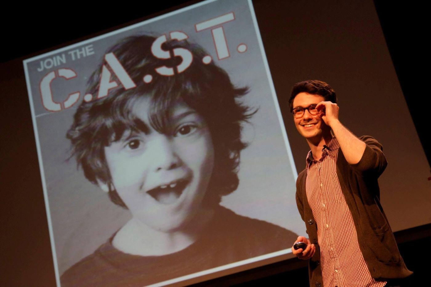 Timothy Doner on stage, a hand raised to the frame of his glasses, a large screen behind him showing a photo of him as a child and the words JOIN THE C.A.S.T.